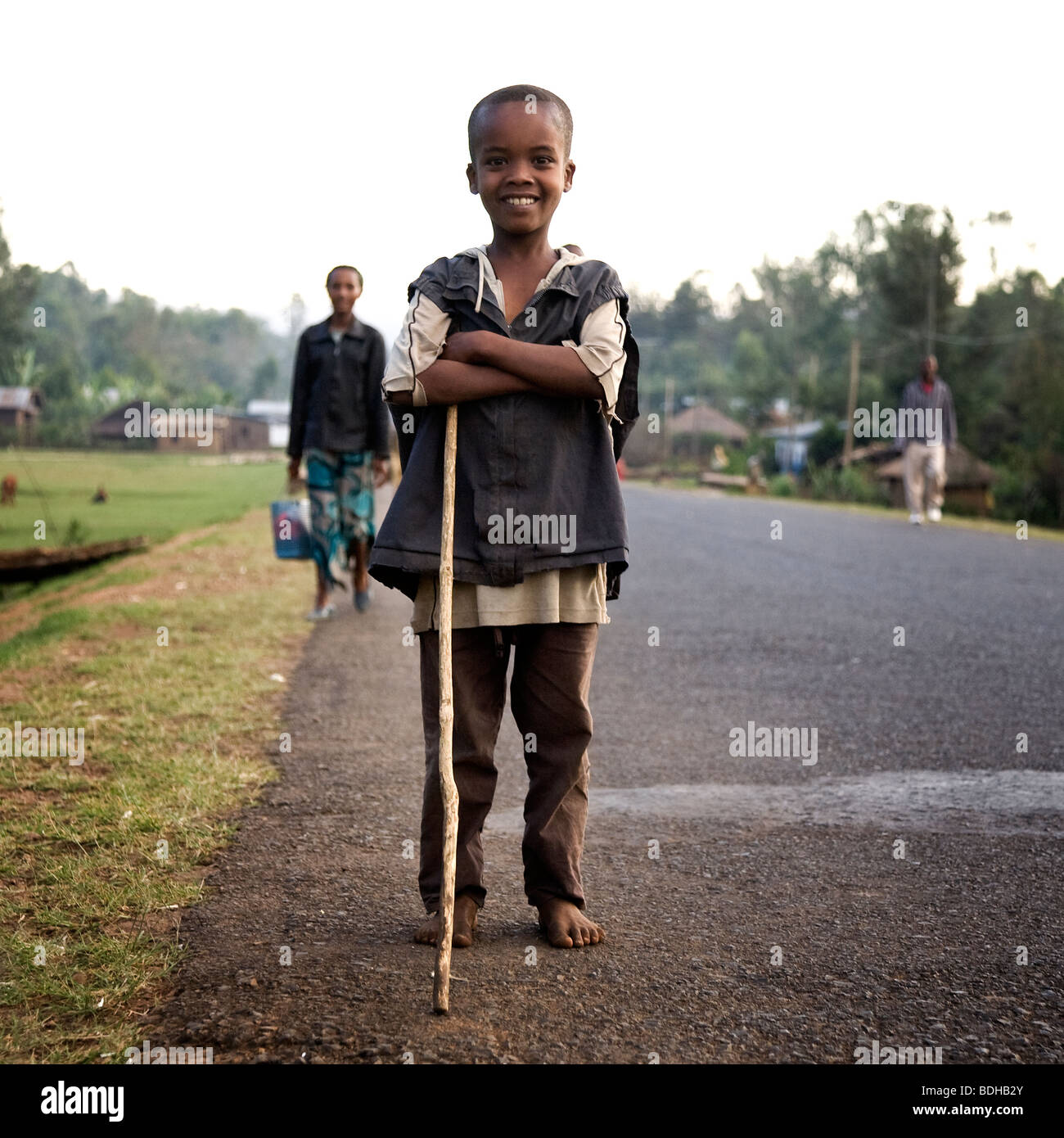 Ein Junge mit einem Gehstock posiert am Rande einer asphaltierten Straße mit Dorfbewohnern zu Fuß die Straße hinter ihm. Stockfoto