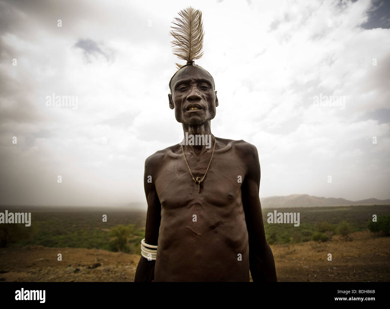 Ein älterer Mann in traditioneller Kleidung und Körper Dekoration stellt vor der krassen Wüstenlandschaft und bewölktem Himmel. Stockfoto