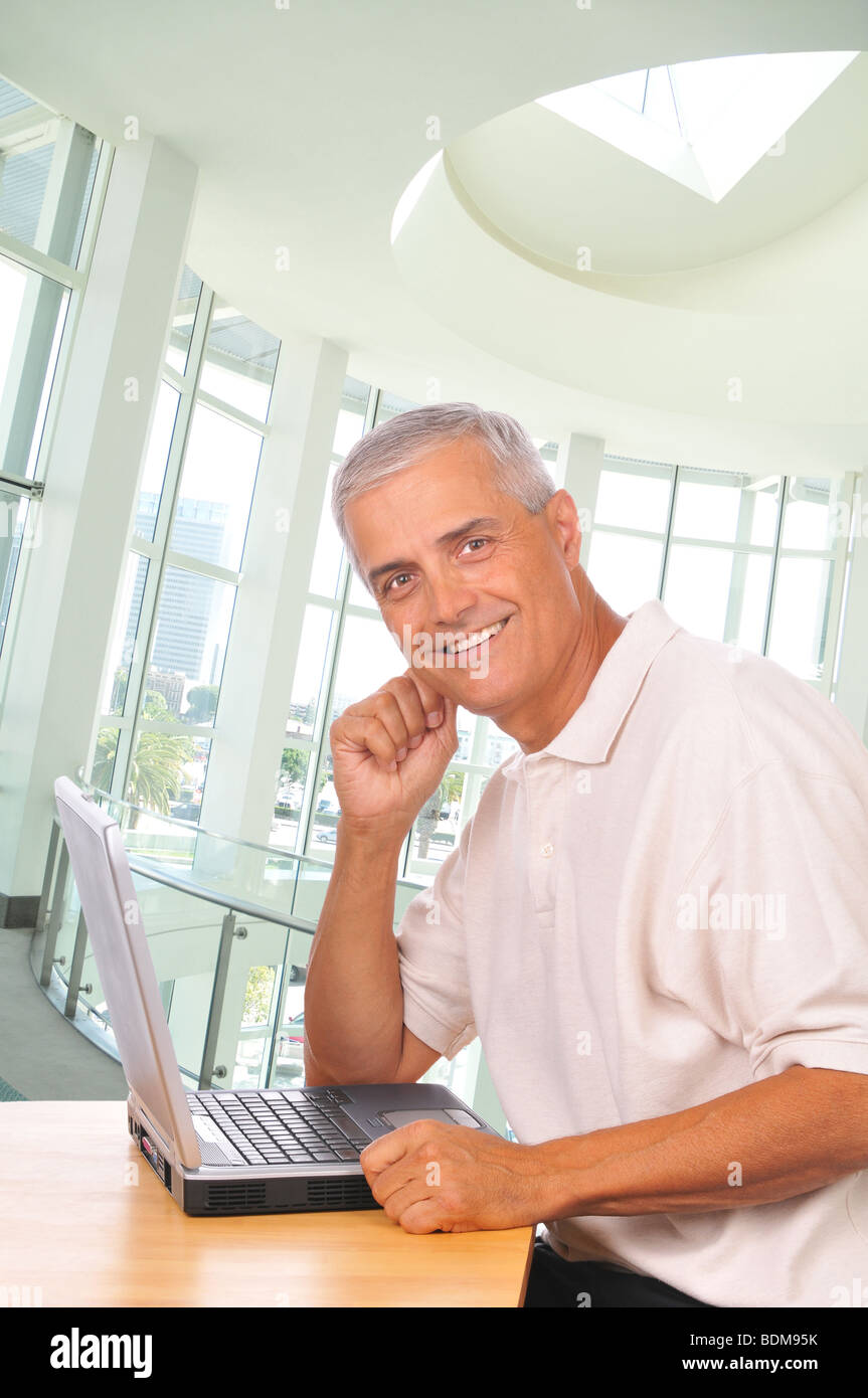 Lächelnd, reifer Geschäftsmann sitzt am Computer gesehen von der Seite in Büroumgebung Stockfoto