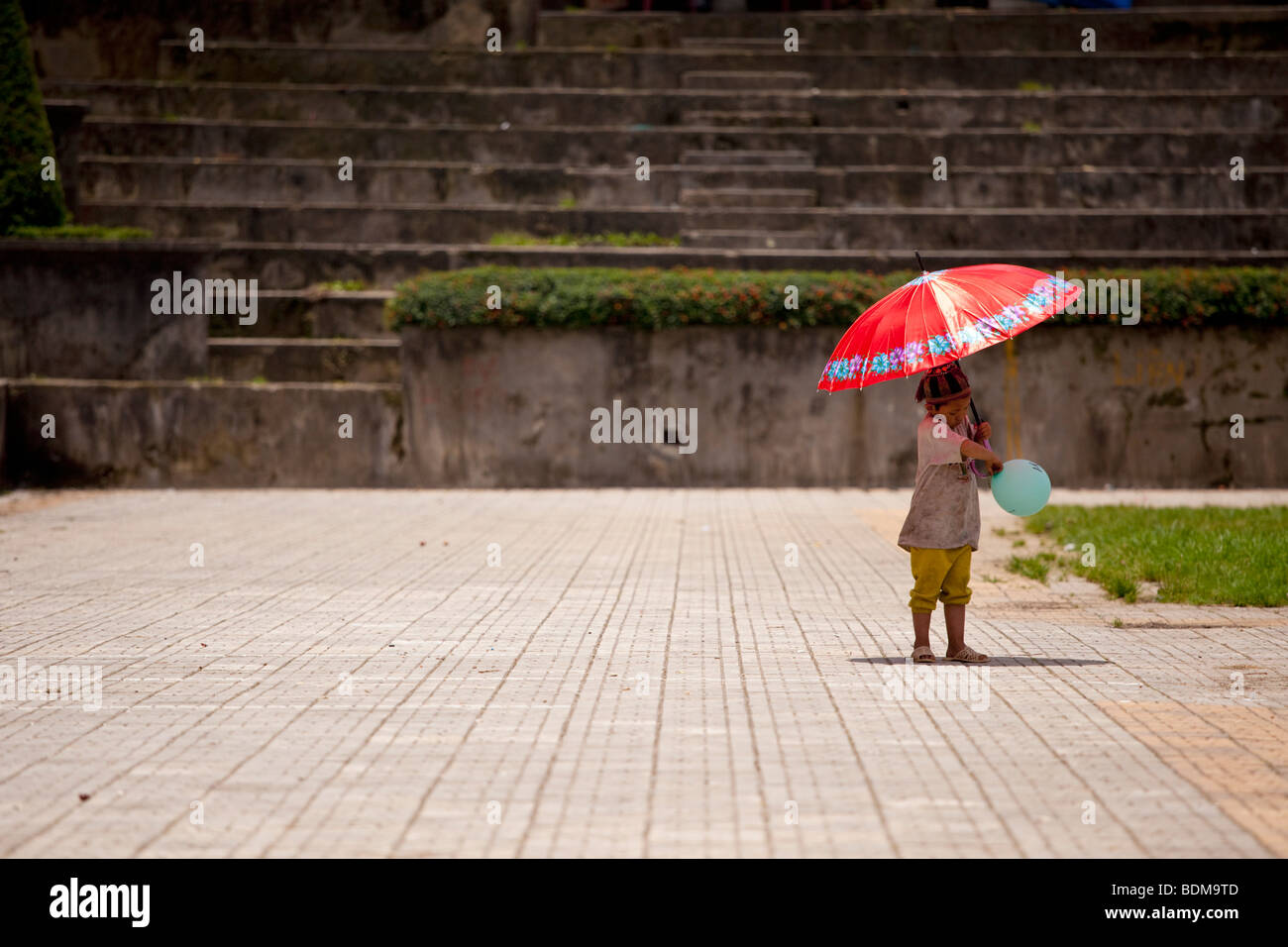 ein einsames Kind spielt mit einem Ballon und leuchtend roten Sonnenschirm Stockfoto