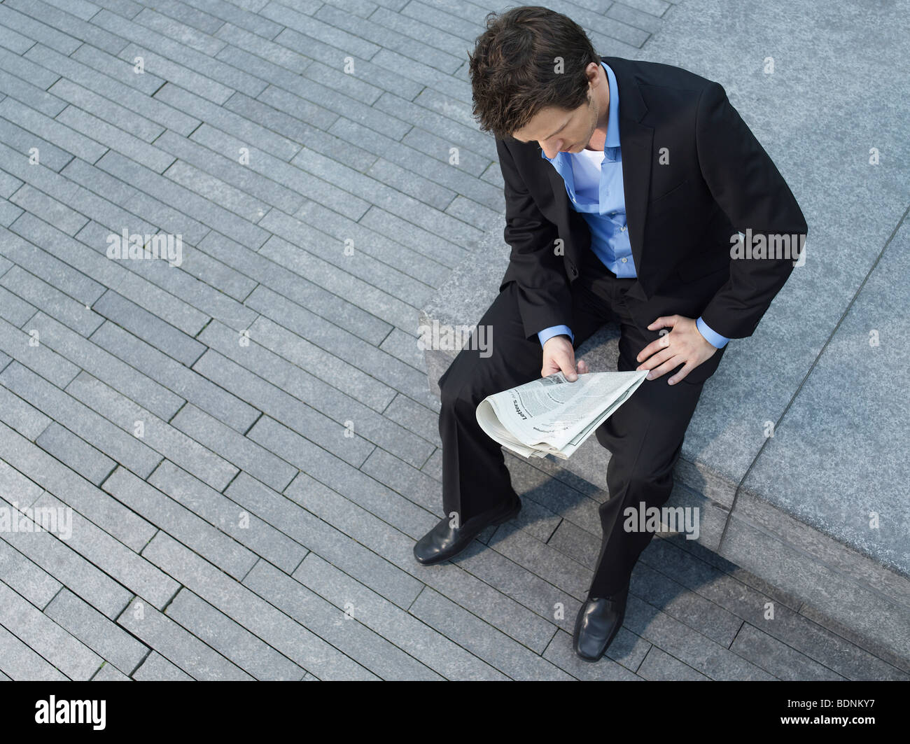 Junger Geschäftsmann sitzt auf der Mauer auf Asphalt, lesen Zeitung, erhöht, Ansicht Stockfoto