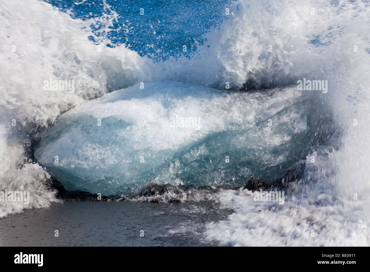Slow-Shutter Temposchuss einer Welle brechen gegen einen Eisberg auf einem vulkanischen schwarzen Sandstrand in Island. Stockfoto