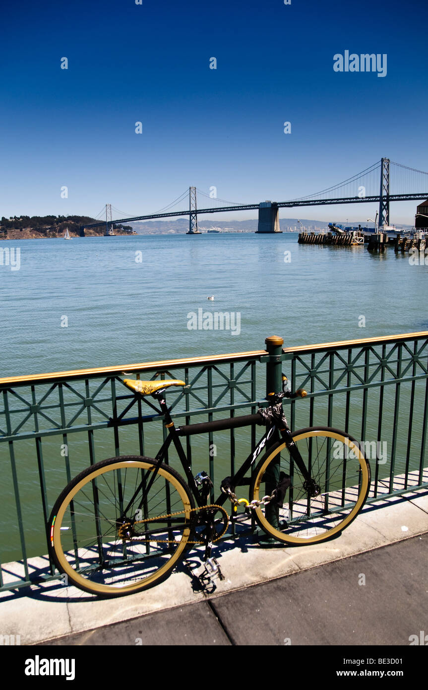 San Francisco Oakland Bay Bridge Bicycle Waterfront San Francisco California // SAN FRANCISCO, Kalifornien — Ein schwarzes Fahrrad mit goldenen Felgen parkt entlang der Uferpromenade, mit der berühmten San Francisco-Oakland Bay Bridge, die sich über die San Francisco Bay erstreckt. Diese große Hängebrücke verbindet San Francisco mit Oakland und anderen Städten der East Bay. Die Brücke erstreckt sich etwa 7,2 Kilometer (4,5 Meilen) über das Wasser. Der klare blaue Himmel und das ruhige Wasser der Bucht von San Francisco bilden eine malerische Kulisse für diese urbane Szene. Diese Ufergegend ist weithin bekannt als ein beliebtes de Stockfoto