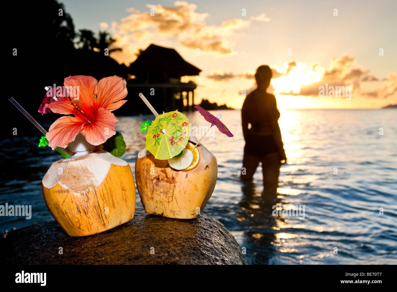 Zwei Kokosnüsse mit Cocktails und Dekorationen auf einem Granitfelsen bei Sonnenuntergang, in den Rücken, die, den eine Frau in das Meer, Seychellen steht Stockfoto