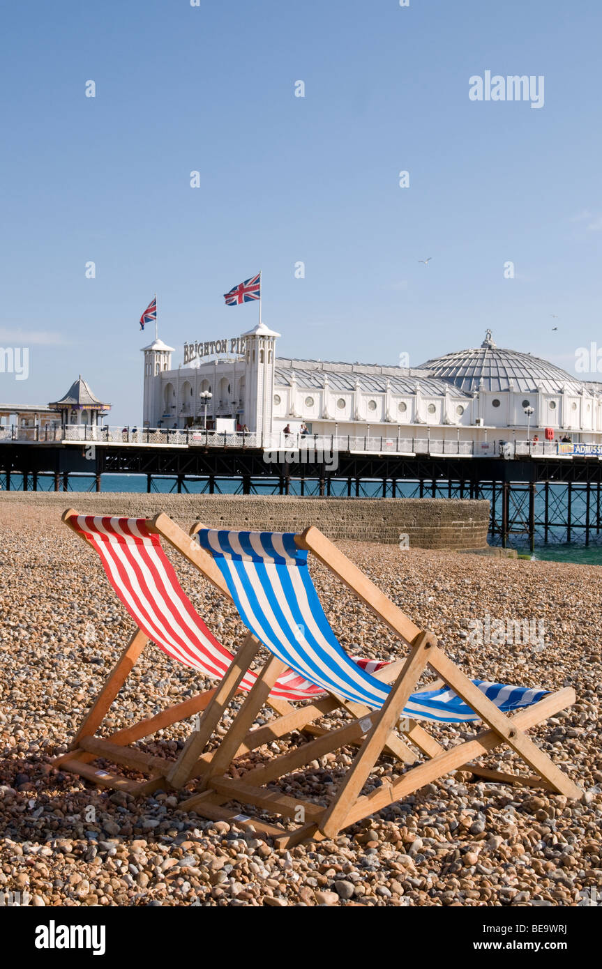 Zwei Liegestühle neben einander am Strand von Brighton, mit dem Palace Pier im Hintergrund. Stockfoto