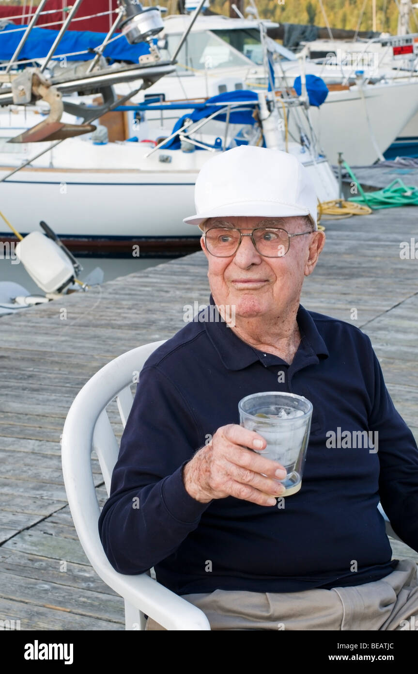 Ein gut aussehender älterer Mann hält einen Drink beim Sitzen auf einem Stuhl auf einer Marina Dock im pazifischen Nordwesten. Stockfoto