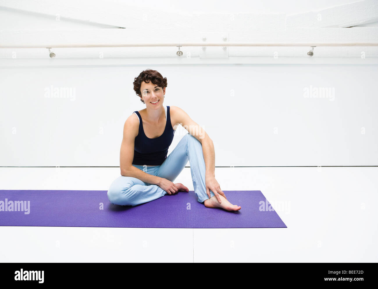 Ein Porträt einer lächelnden Frau sitzt in einem weißen Raum auf einer Yogamatte. Stockfoto