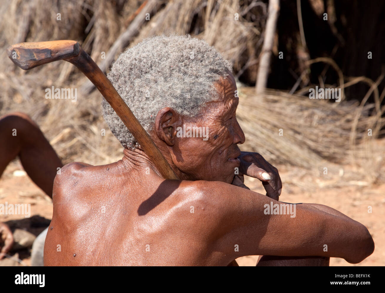 San-Dorf. Einen grauen Haaren alten San sitzt Man im Dorf, hält er einen Holzknüppel. Stockfoto