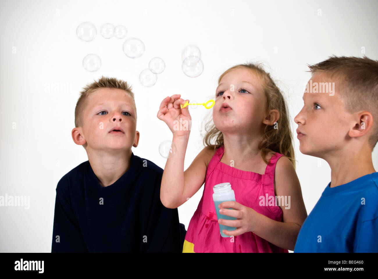 Ein kleines Mädchen bläst Seifenblasen durch einen seifigen Zauberstab während ihr Aussehen Brüder auf. Stockfoto