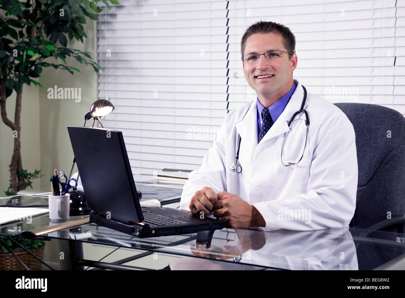 Eine lächelnde junge Ärztin sitzt an seinem Schreibtisch arbeitet an seinem Laptop. Stockfoto