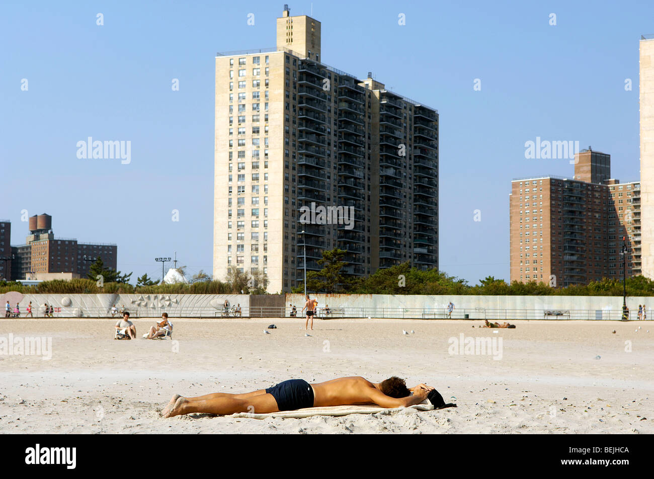Ein Mann schläft auf den Sand am Strand von Coney Island, Brooklyn, New York. Hochhaus-Wohnprojekte sind im Hintergrund abgebildet. Stockfoto