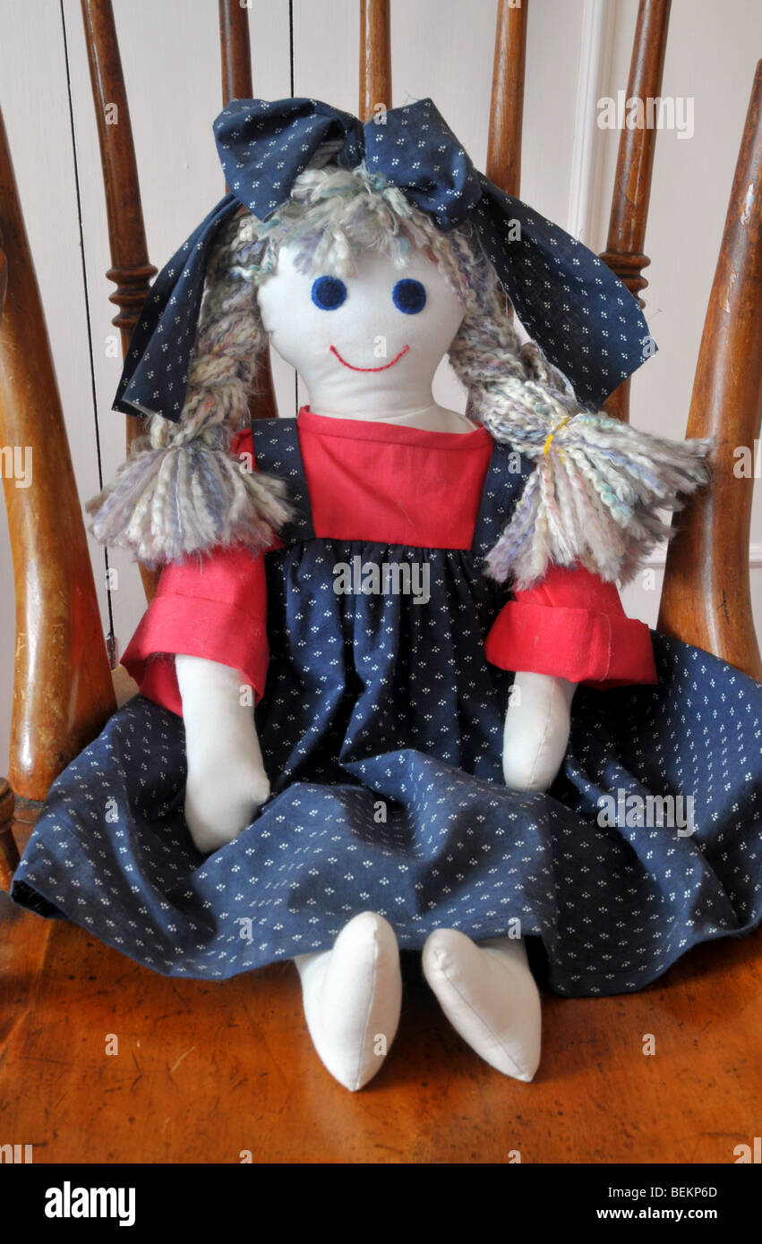 Eine hausgemachte Stoff Puppe gekleidet in blaue und rote Schürze mit einer Schleife im Haar, sitzt auf einem Holzstuhl. Stockfoto