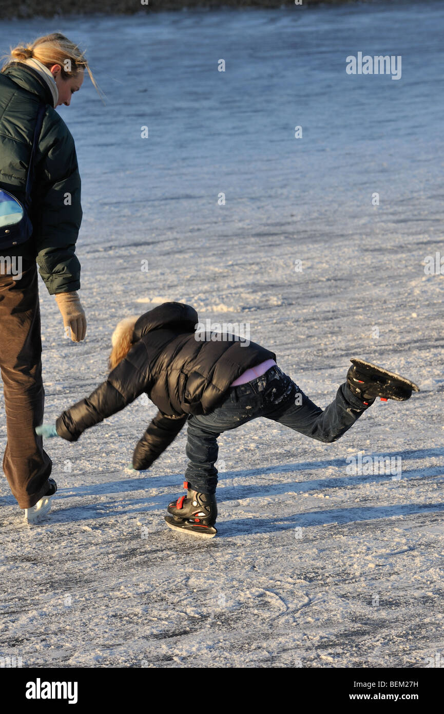 Kind fällt neben Mutter während Sie auf dem Eis am zugefrorenen Kanal im Winter Schlittschuh laufen lernen Stockfoto