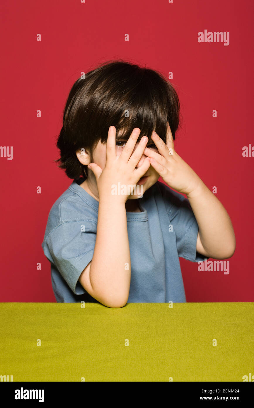 Kleiner Junge bedeckte Gesicht mit den Händen, durch die Finger in die Kamera guckend Stockfoto