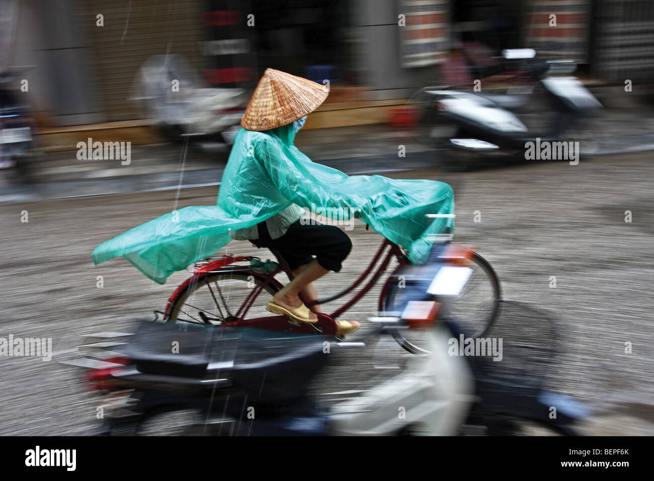 Schwenken die Straßenszene. Frau mit traditionellen konischen Hut mit ihrem Fahrrad unter dem Regen in Hanoi Hauptstadt von Vietnam, Asien Stockfoto