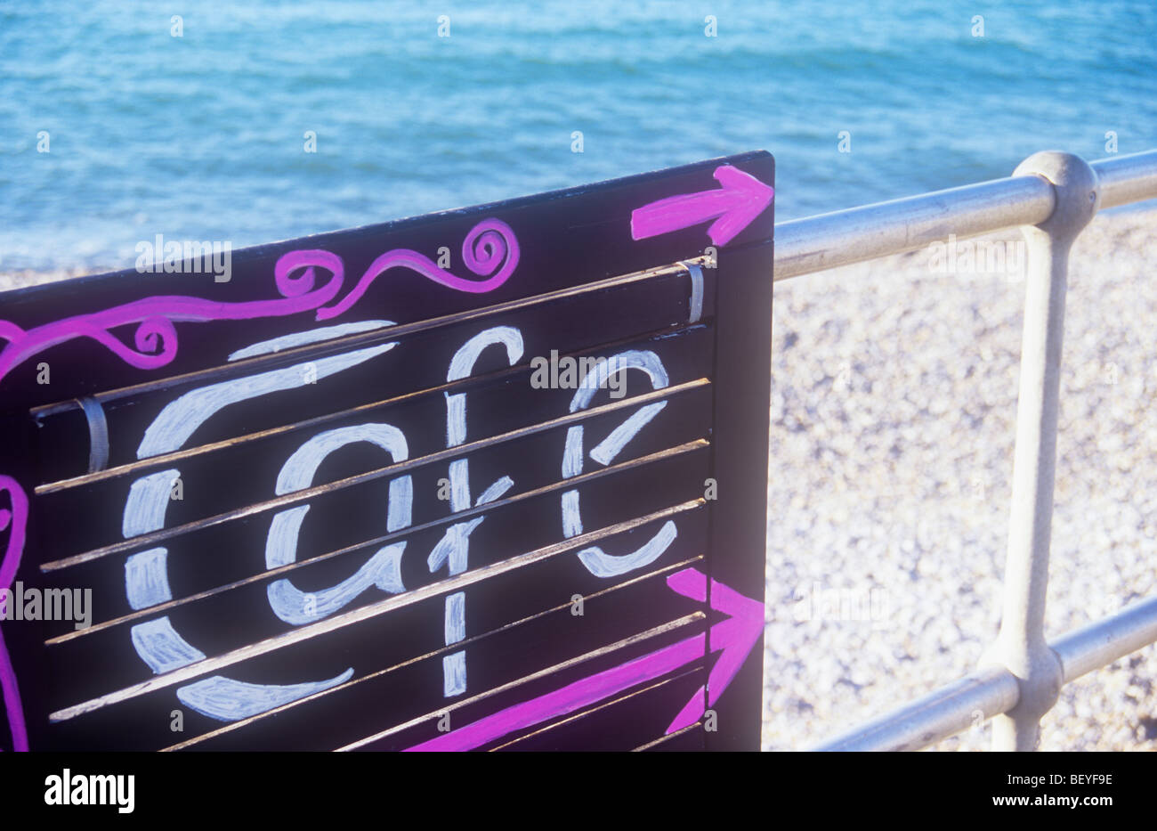 Handgemalte jaunty anmelden, weiß und pink auf schwarz besagt Cafe und fest mit Geländer mit Kiesstrand und blaues Meer hinter Stockfoto