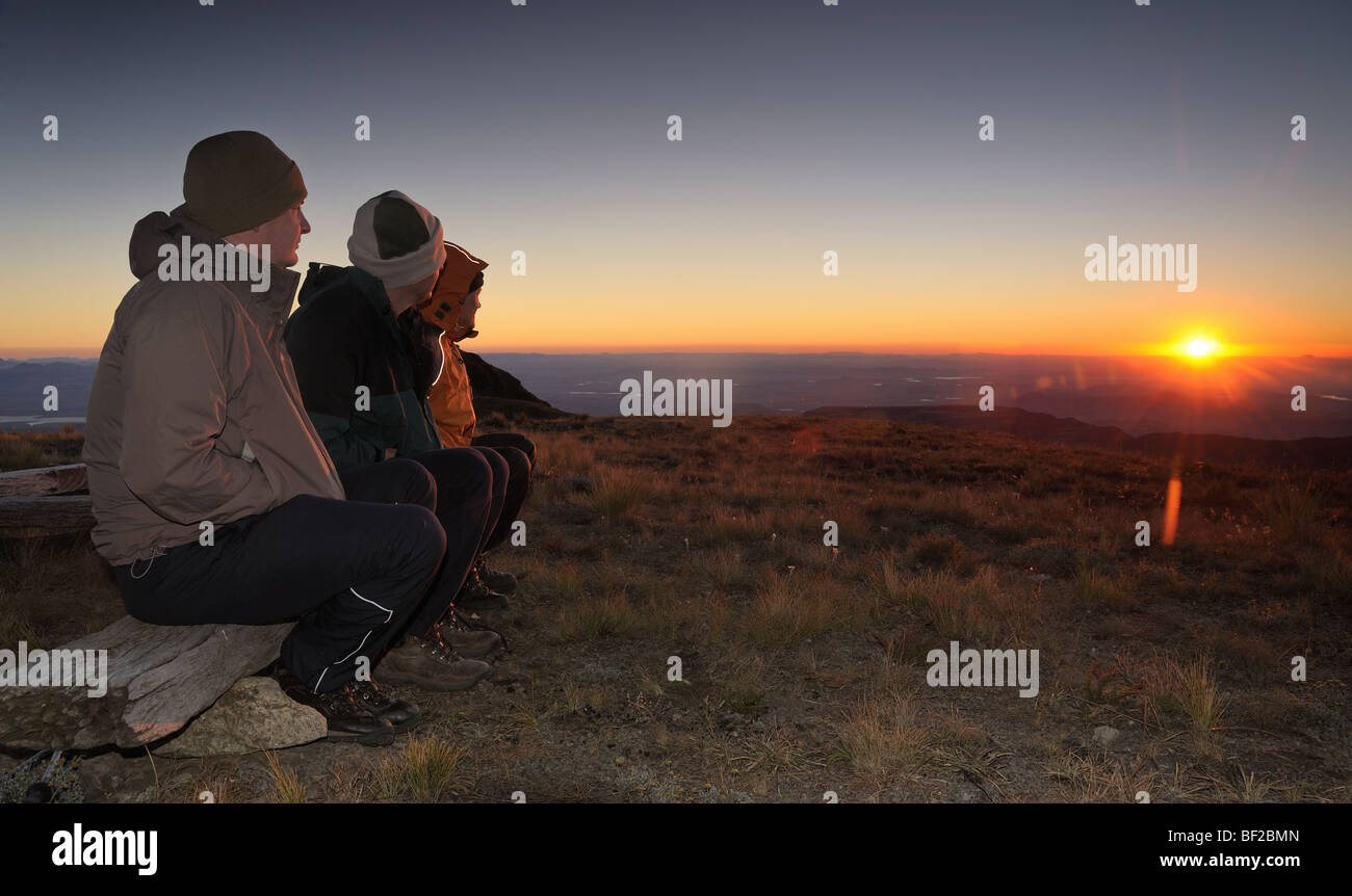 Drei Wanderer gekleidet in warme Kleidung, die den Sonnenaufgang über dem Horizont, Cathedral Peak Drakensberg Ukahlamba nationale Par Stockfoto