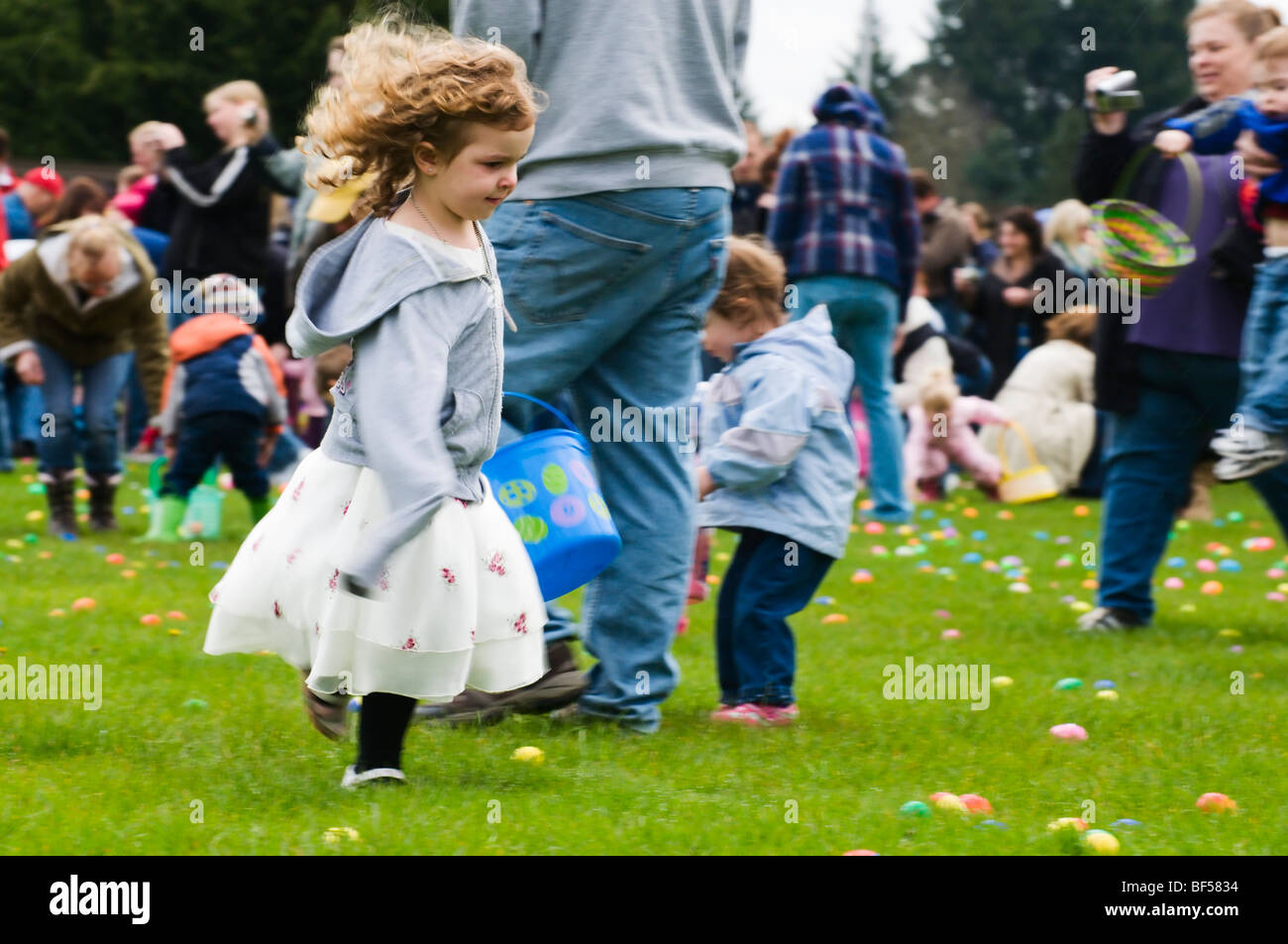 Ein Liebling kleines Mädchen läuft durch den Rasen Plastikeier zu sammeln, wie sie in einer Gemeinschaft Ostereiersuche teilnimmt. Stockfoto