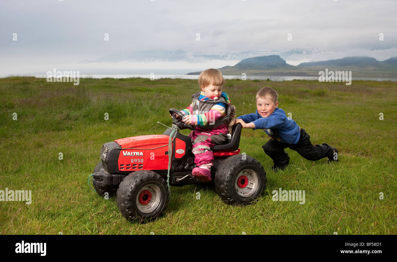 Kleiner Junge schob seine jüngere Schwester auf Spielzeug-Traktor, Eskifjordur, Island Stockfoto