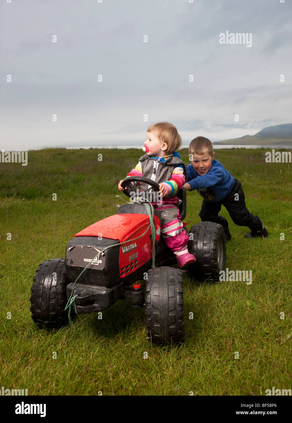 Kleiner Junge schob seine jüngere Schwester auf Spielzeug-Traktor, Eskifjordur, Island Stockfoto
