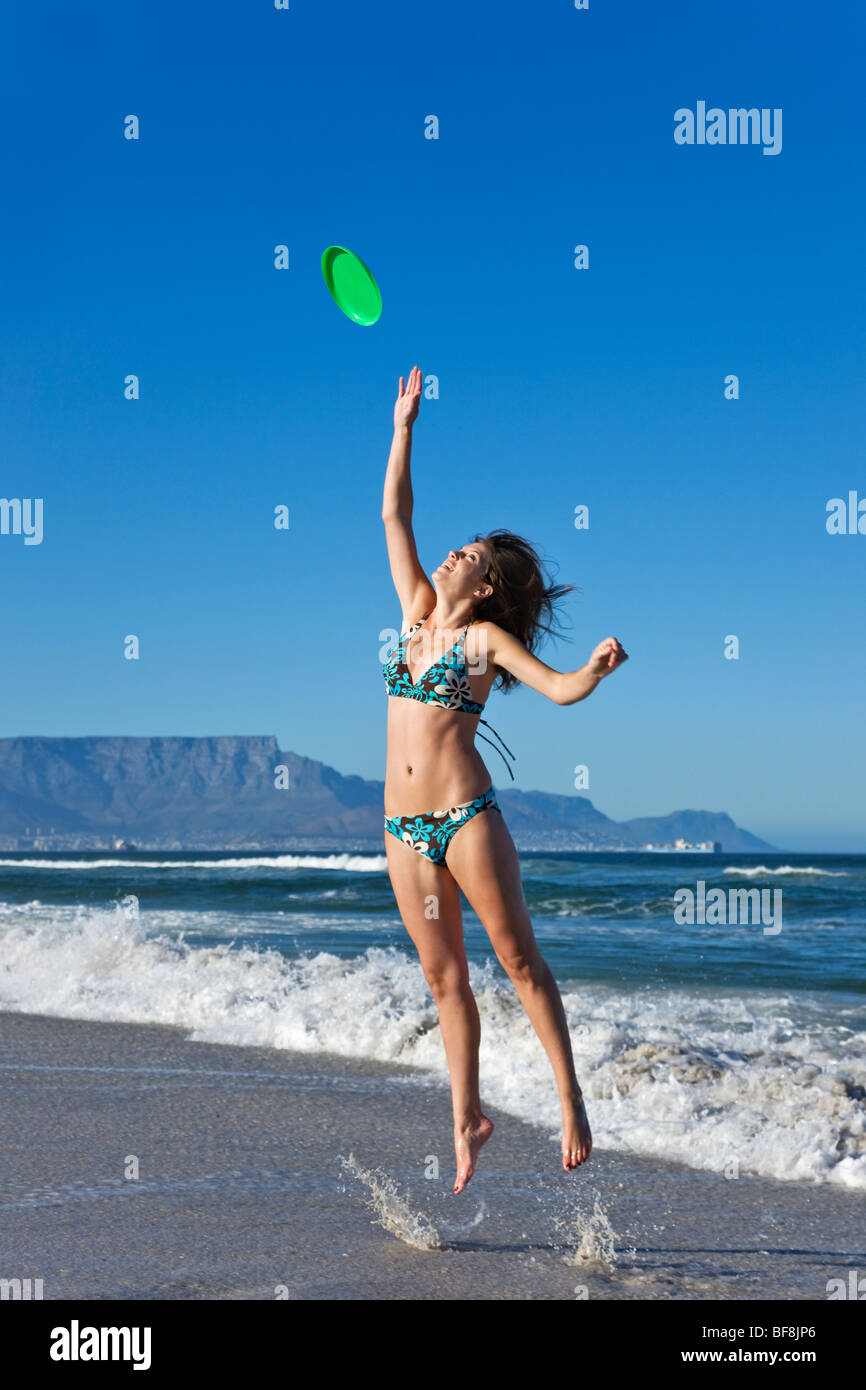 Junge Frau trägt eine Badehose springen, um einen Frisbee in den seichten Wellen zu fangen. Tafelberg im Hintergrund Kapstadt Stockfoto