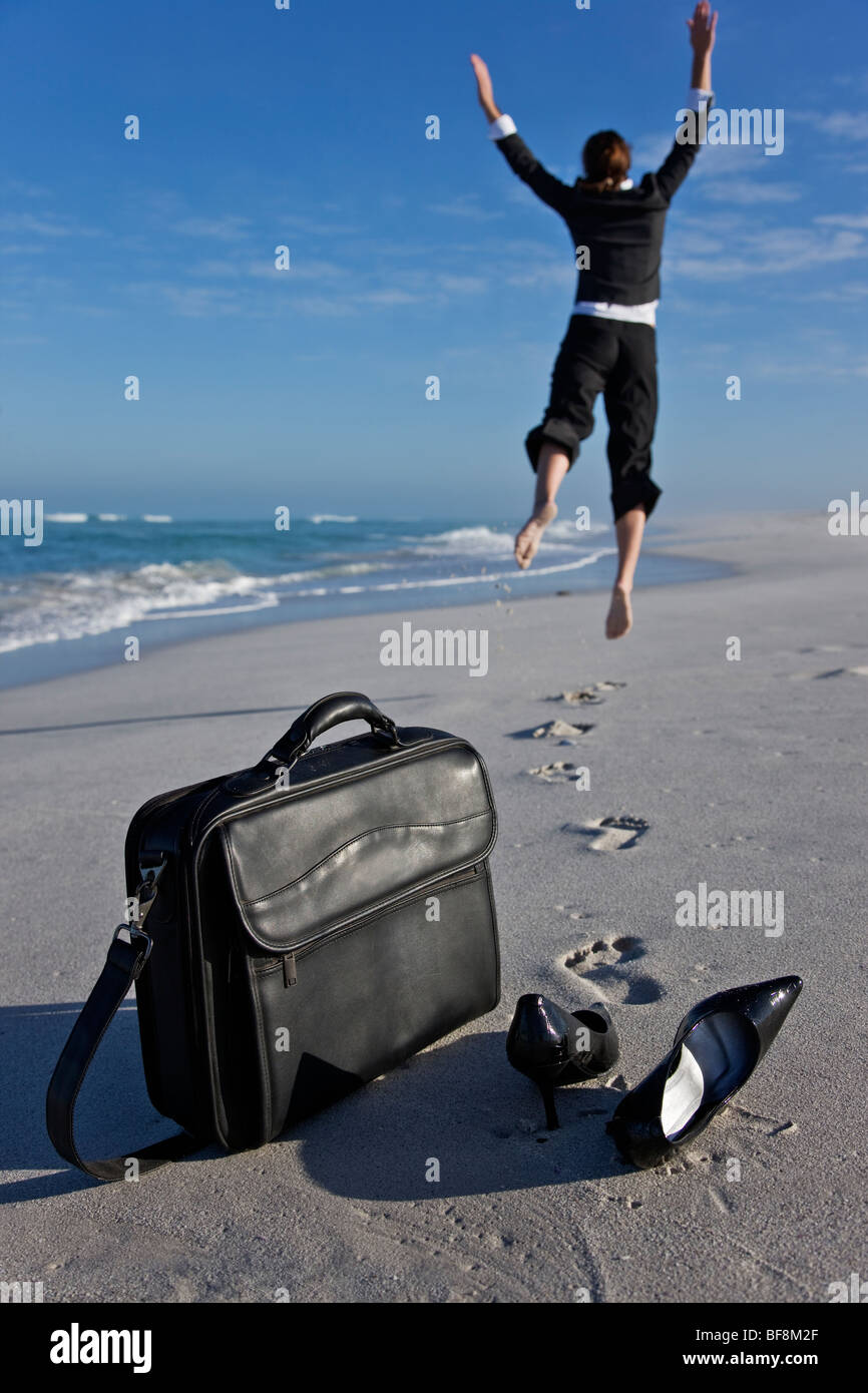 Frau im Büro Kleidung am Strand springen mit Freude und Freiheit. Laptop-Tasche und Schuhe im Vordergrund. Stockfoto