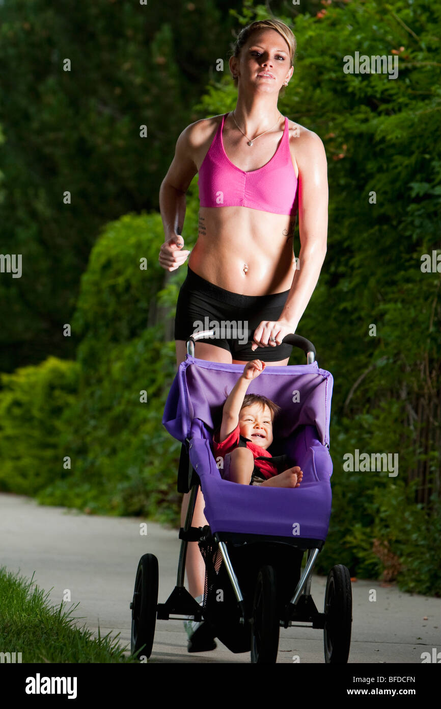 Baby wirft Faust lächelnd in jogging-Kinderwagen geschoben durch eine kantig aussehende, sportliche junge Frau während eines Laufes auf einem suburban Bürgersteig Stockfoto