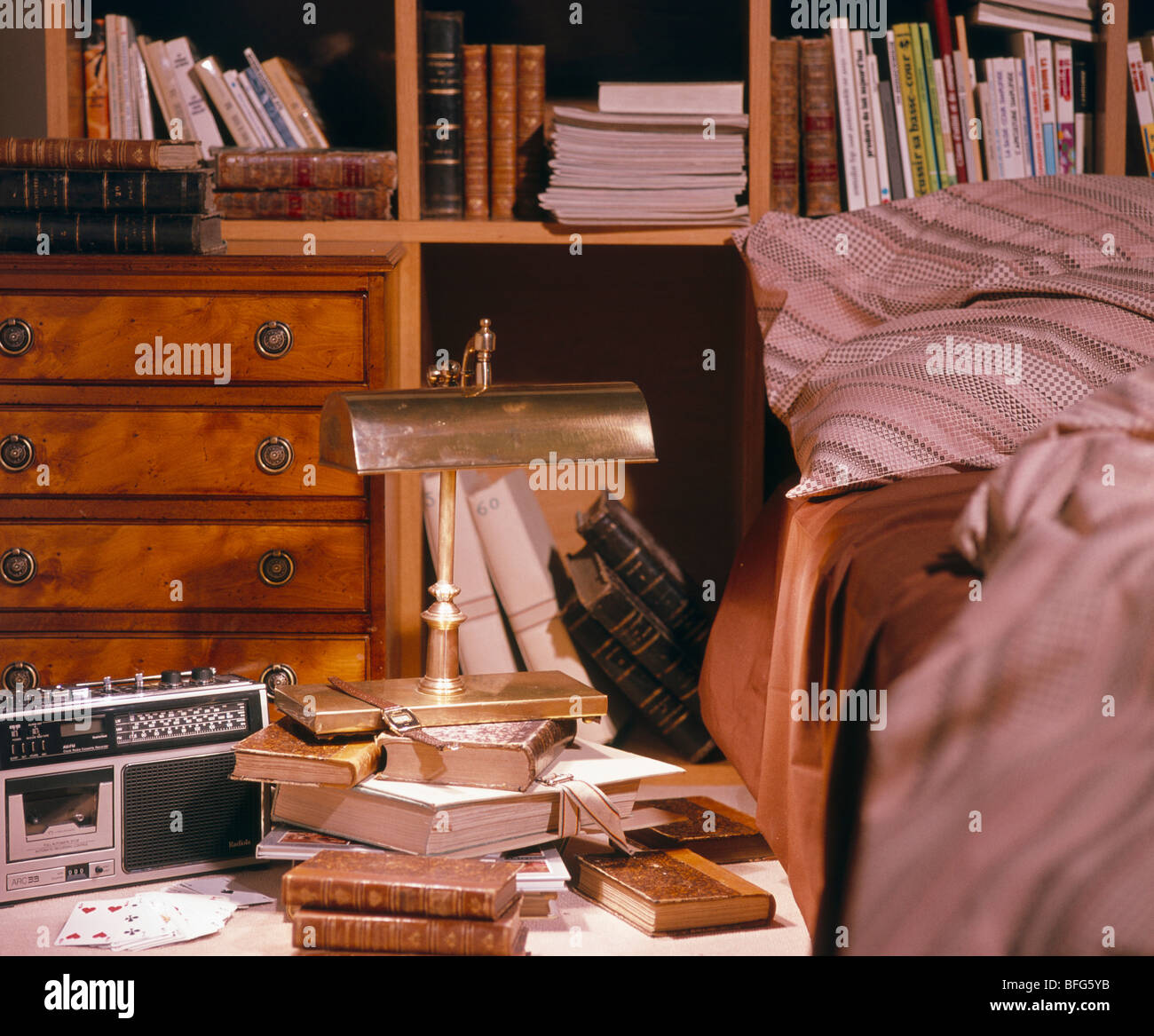 Kleines Radio und antike Messinglampe mit Büchern auf Nachttisch vor Bücherregalen im traditionellen Schlafzimmer Stockfoto