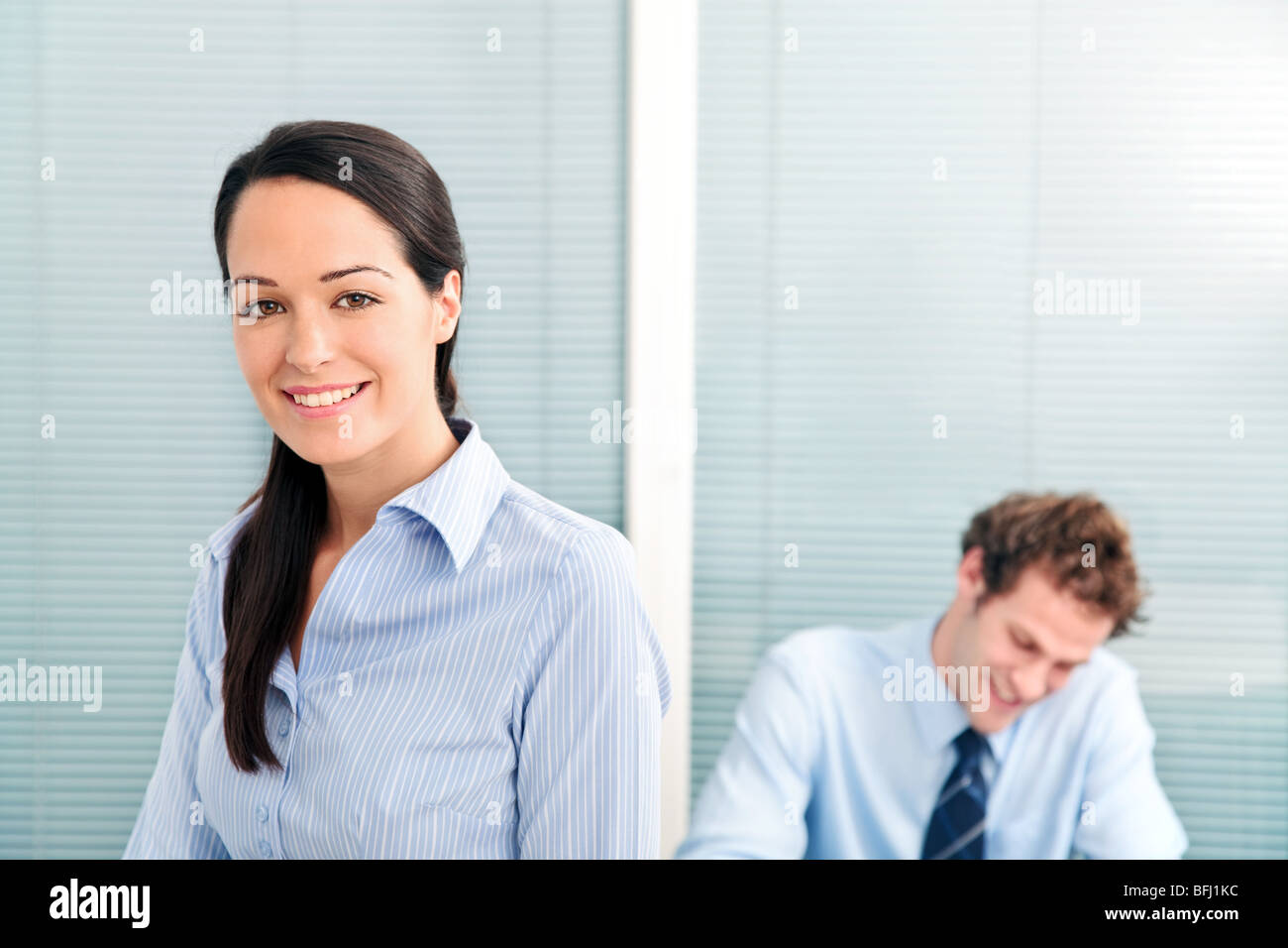 Geschäftsfrau in einem Büro lächelnd zu Kamera, männlicher Kollege im Hintergrund unscharf. Stockfoto