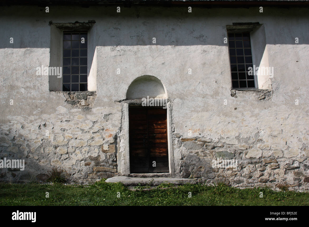 Hölzerne Tür und zwei Fenster eines alten ländlichen Gebäude. Foto von außen. Weiße Wand mit Markierungen der bröckelnden Putz. Schatten auf die Tür. Stockfoto