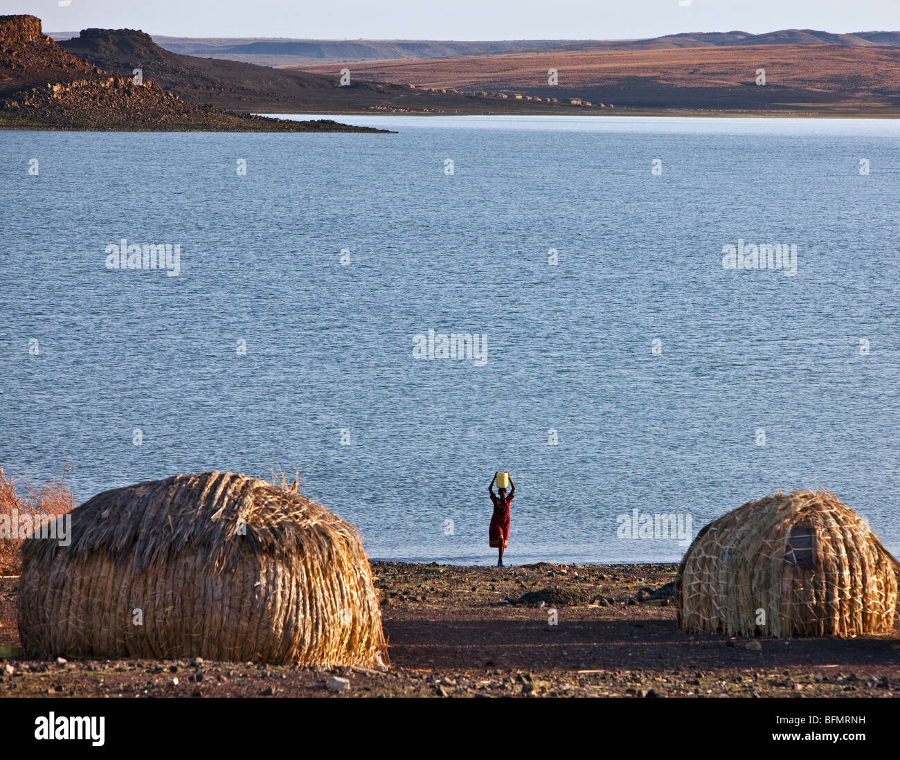 Eine El Molo Frau holt Wasser vom Turkana-See. Typische kuppelförmige El Molo Häuser im Vordergrund sind aus Schilf hergestellt. Stockfoto