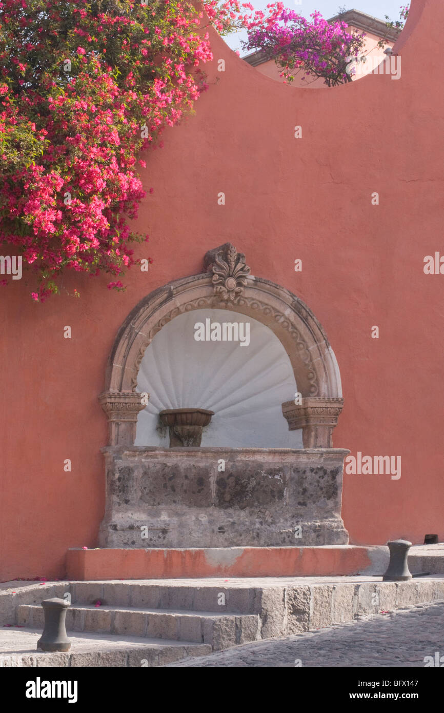 Bogen Sie geformte Brunnen in einem Adobe Terrakotta Wand, rosa Bougainvillea. Graue Treppe führt diagonal von Straße mit Kopfsteinpflaster. Stockfoto