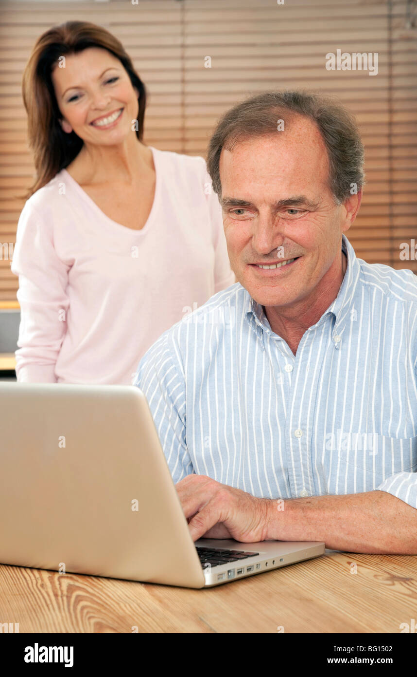 Lächelnde Frau schaut Ehemann oder Partner, wie er einen silbernen Laptop-Computer am Küchentisch benutzt Stockfoto
