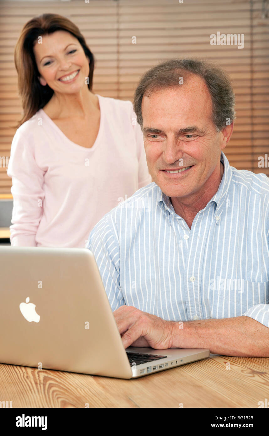 Lächelnde Frau schaut Ehemann oder Partner, wie er einen Mac-Laptop-Computer am Küchentisch benutzt Stockfoto