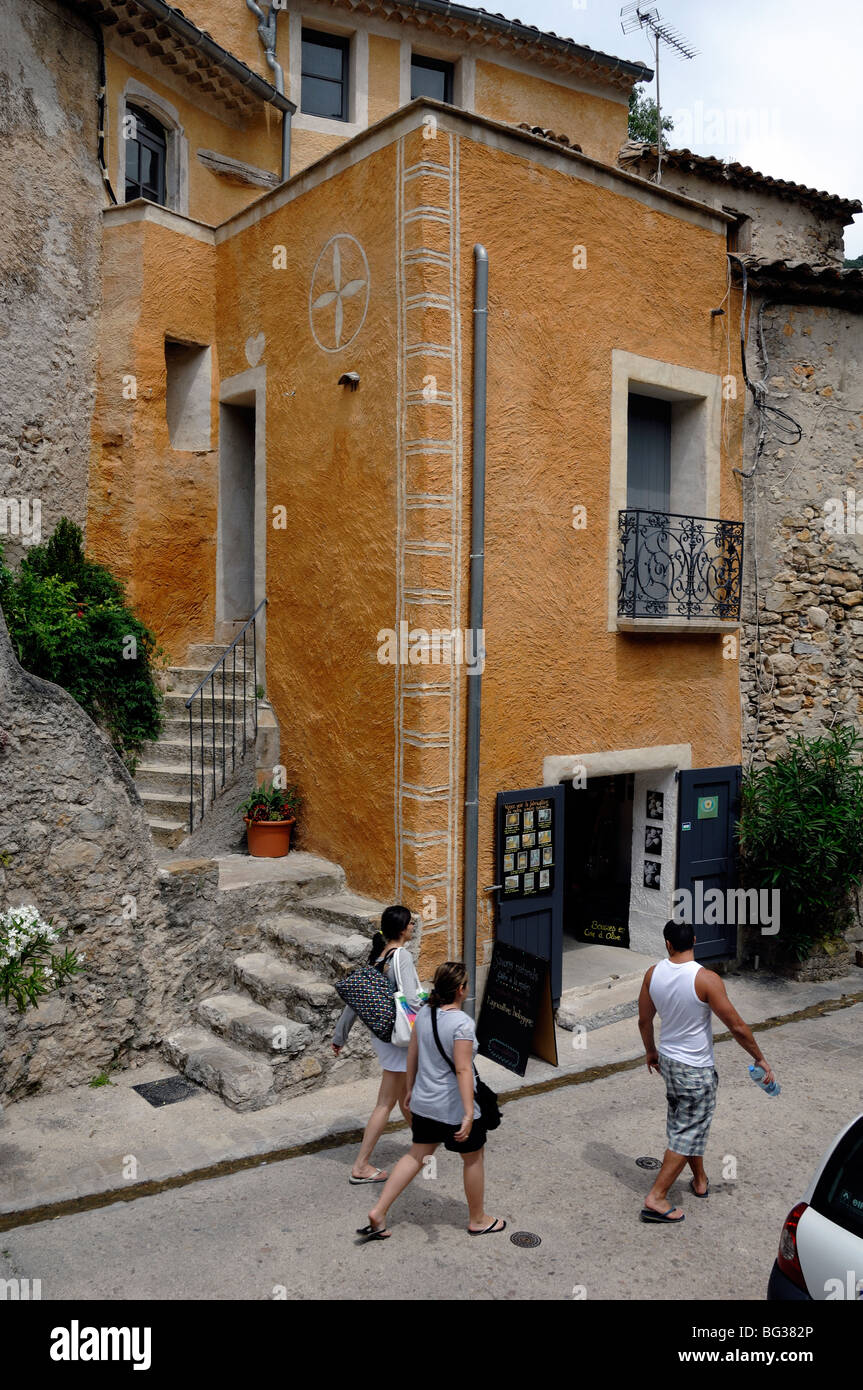 Ockerfarbenes Haus, Boutique & drei Touristen, die die Hauptstraße entlang gehen, Saint-Guilhem le Désert, Hérault, Frankreich Stockfoto