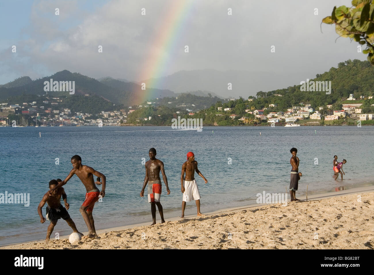 Jugendliche, die Fußball spielen in der Sonne auf einer White Coral Beach mit einem Regenbogen hinter ihnen. Grand Anse Beach, Grenada, Karibik. Stockfoto