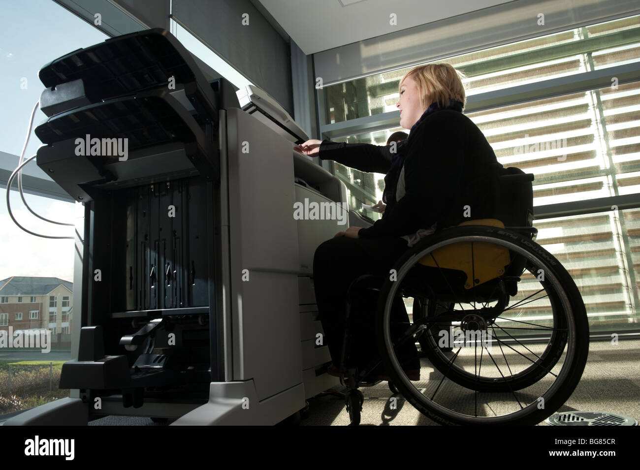 Eine junge behinderte Frau in einem Rollstuhl mit einem Kopierer Maschine in einem modernen Büro, UK Stockfoto