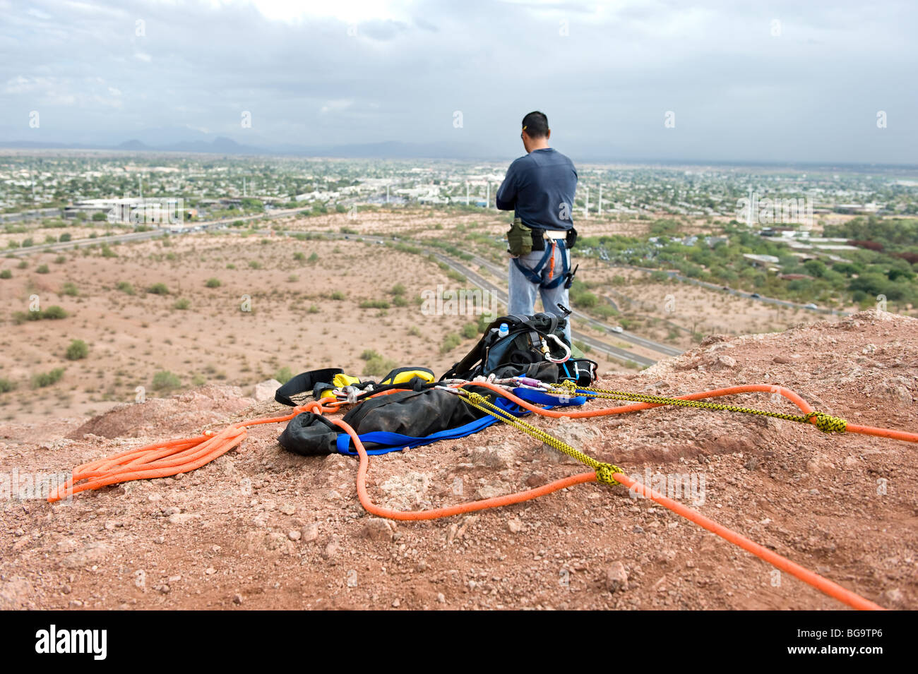 Ein Kletterer, mit Blick auf eine weit entfernte Stadt bereitet sich auf einem steilen und hohen Berggipfel Abseilen und denkt über seinen Plan. Stockfoto