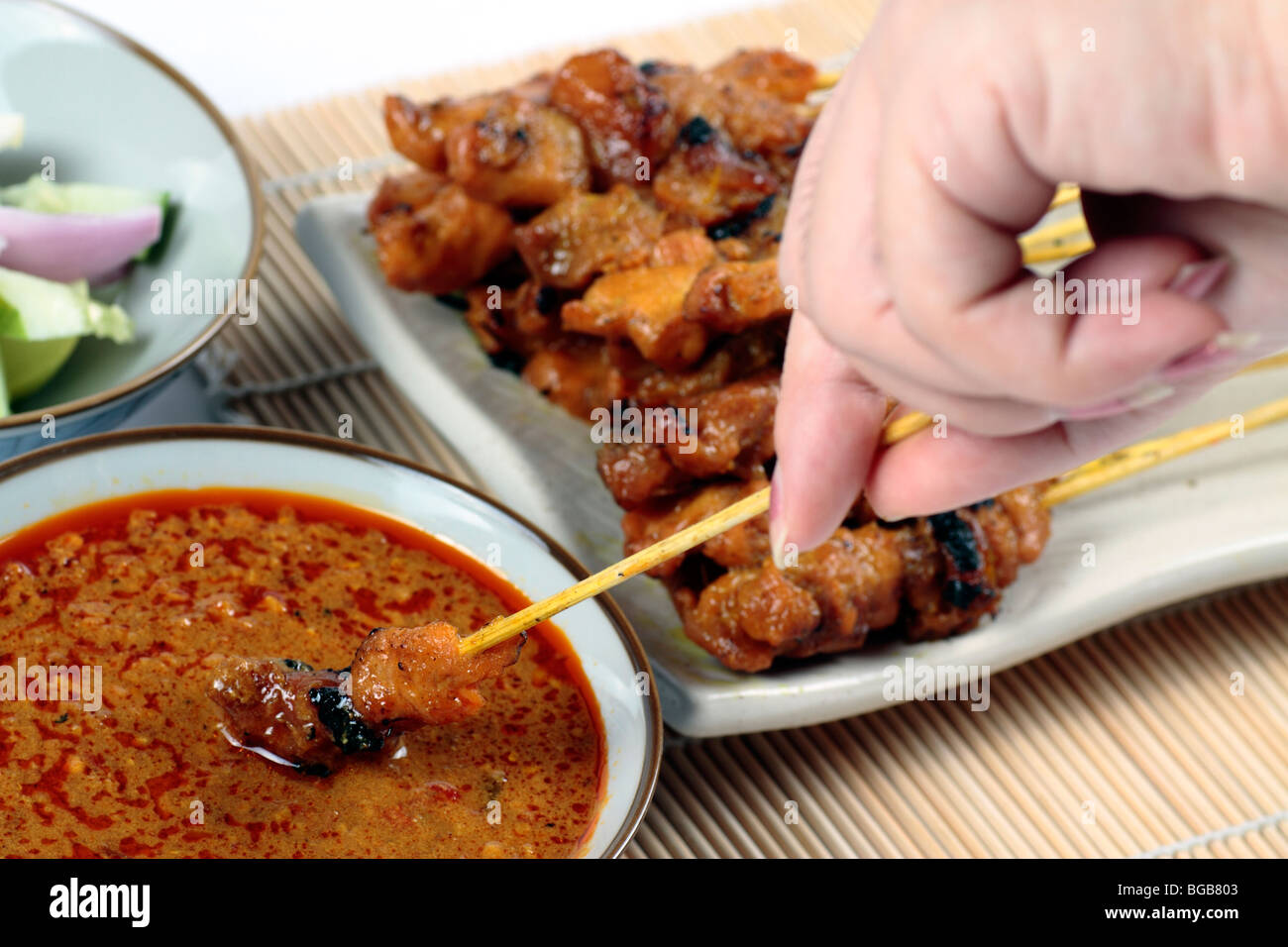 Die Hand einer Frau ein Stück Hühnchen Satay eintauchen, in eine Schüssel mit Erdnuss-Sauce mit mehr Satay im Hintergrund Stockfoto