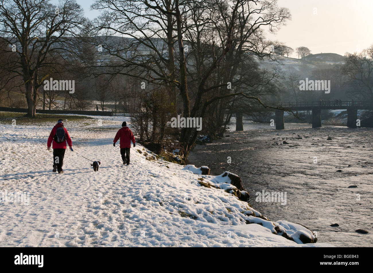 Zwei Wanderer mit ihrem Hund spazieren im Schnee entlang dem Fluss Wharf in der Nähe von Bolton Abbey, North Yorkshire Stockfoto