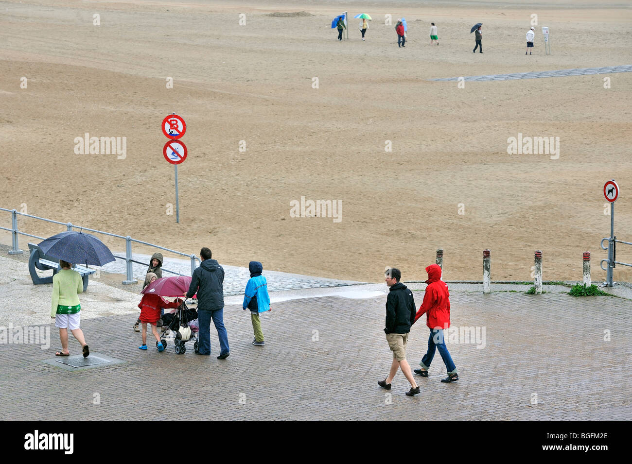 Touristen mit Sonnenschirmen zu Fuß über den Deich entlang Strand an einem regnerischen Tag in den Sommerferien an der belgischen Küste Stockfoto