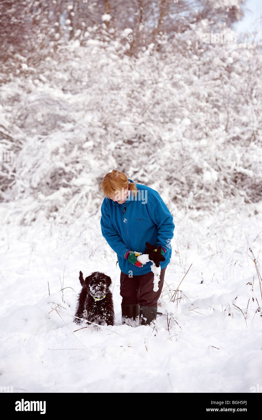 Dame in blauem Vlies wischt den Schnee von einem Hundespielzeug, während ihr tief verschneiten Labradoodle Welpen beobachtet und wartet Stockfoto