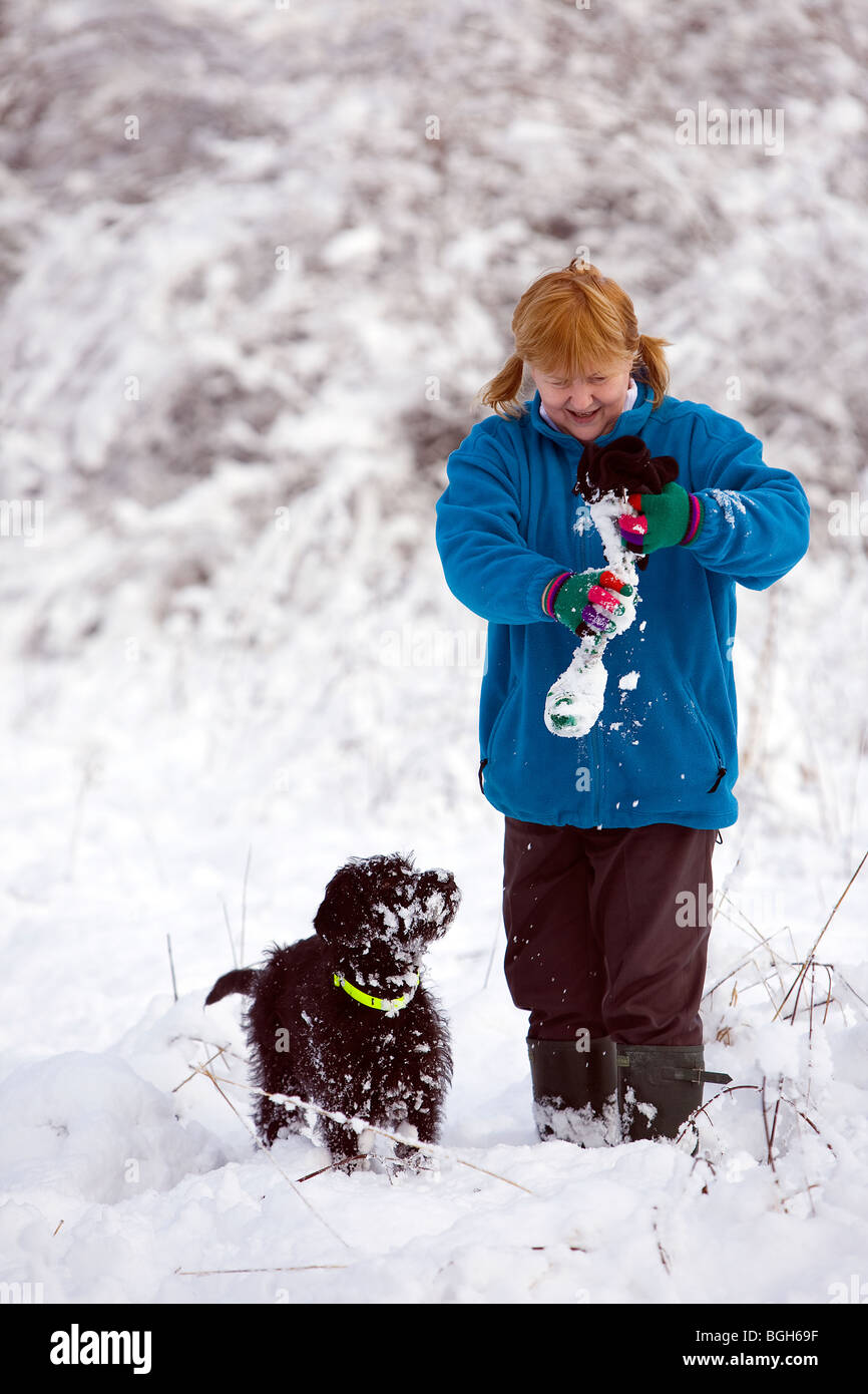 Dame in blauem Vlies wischt den Schnee von einem Hundespielzeug, während ihr tief verschneiten Labradoodle Welpen beobachtet und wartet Stockfoto
