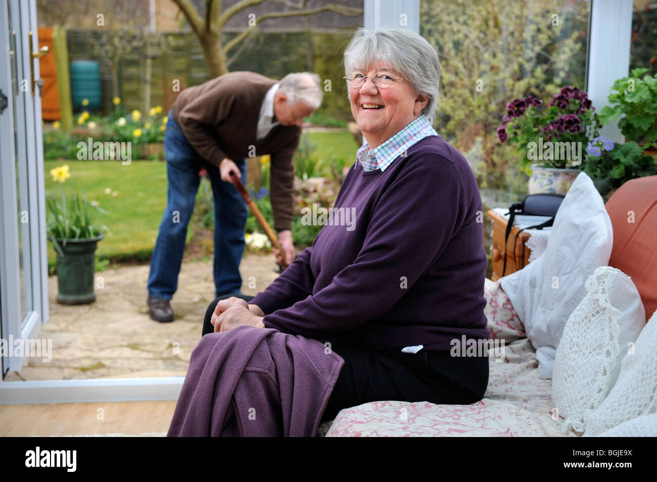 Eine pensionierte Dame in ihrem Wintergarten während ein Mann das Blumenbeet UK Gabeln Stockfoto