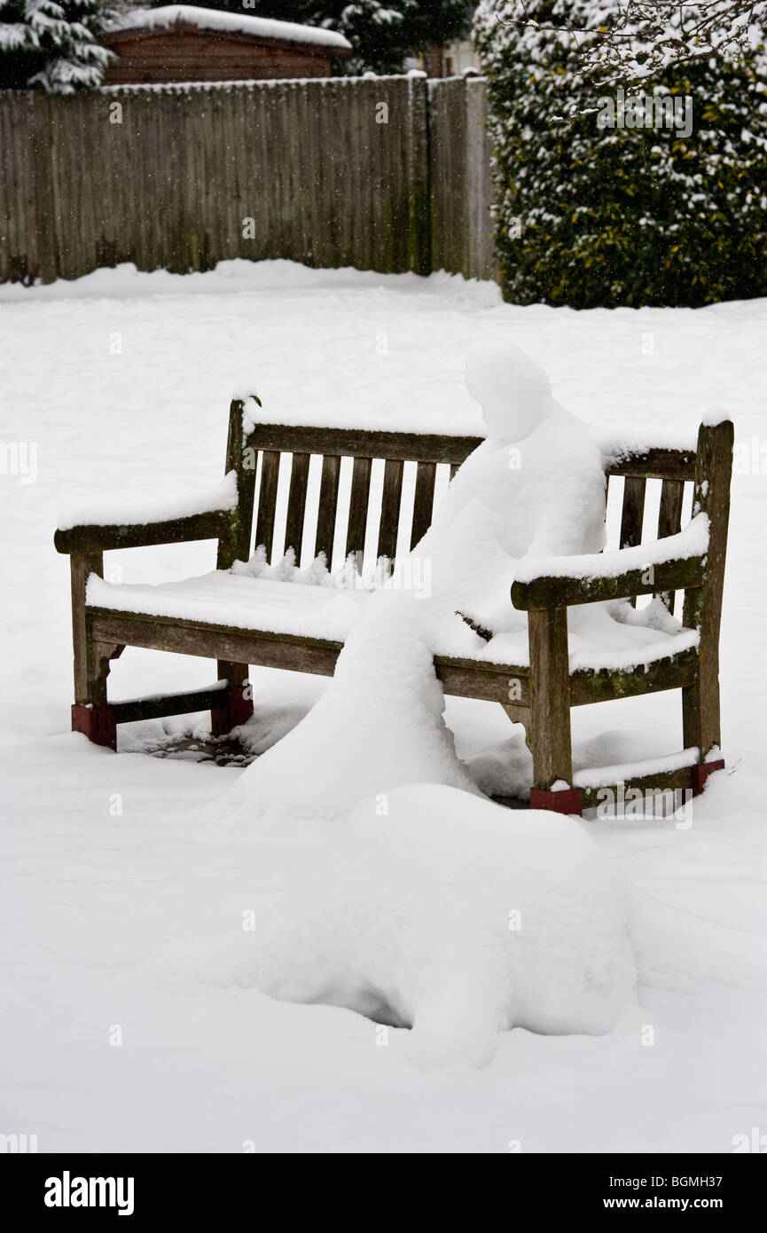 Schneemann auf einer öffentlichen Bank auf einem Dorfplatz im Winterschnee bedeckt. Stockfoto