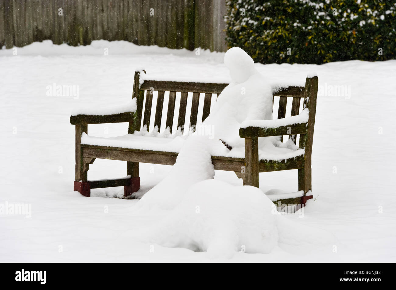 Schneemann auf einer öffentlichen Bank auf einem Dorfplatz im Winterschnee bedeckt. Stockfoto