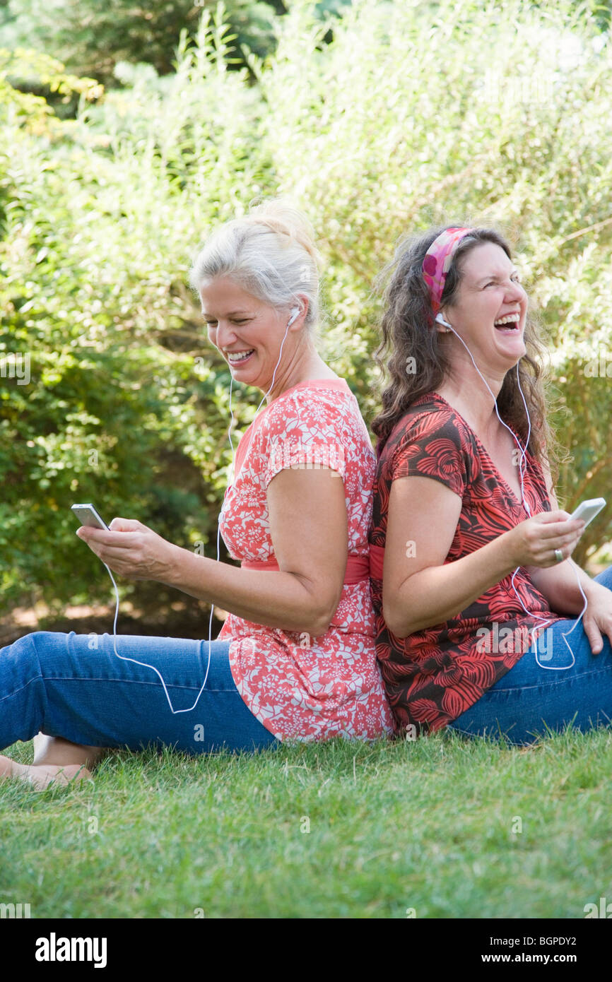 Seitenansicht der zwei Reife Frauen sitzen Rücken an Rücken in einem Park und anhören von MP3-Playern Stockfoto