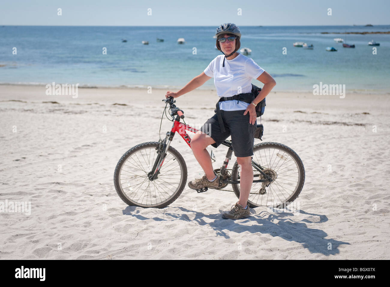 Eine 50 Jahre alte Frau am Strand mit einem Fahrrad mit Fahrradhelm und Sonnenbrillen in Kerity, nahe Penmarc'h, Bretagne, Frankreich Stockfoto