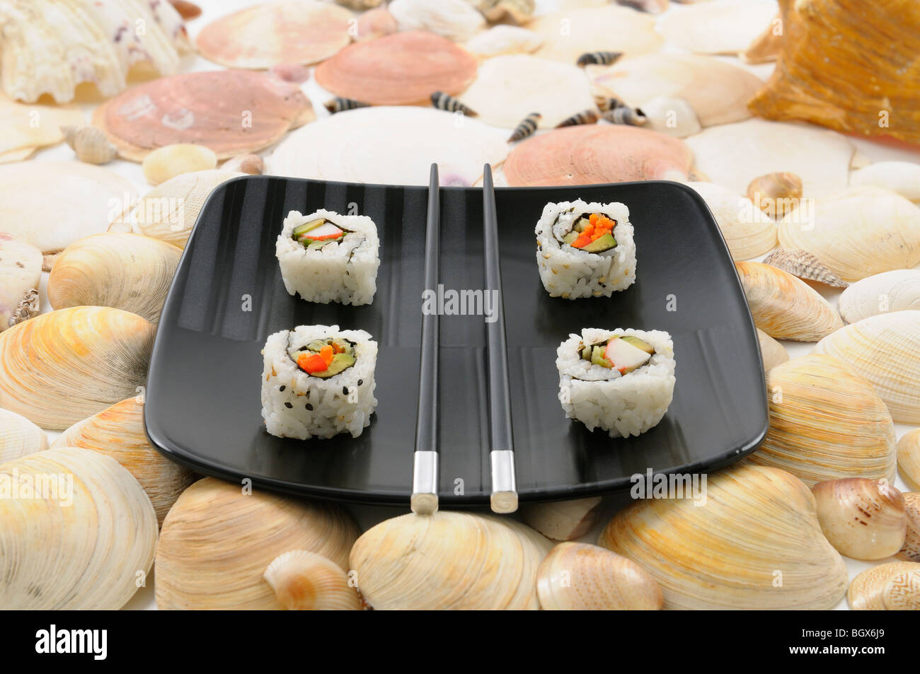 Uramaki Sushi und Stäbchen auf einer schwarzen Platte umgeben von Muscheln Stockfoto