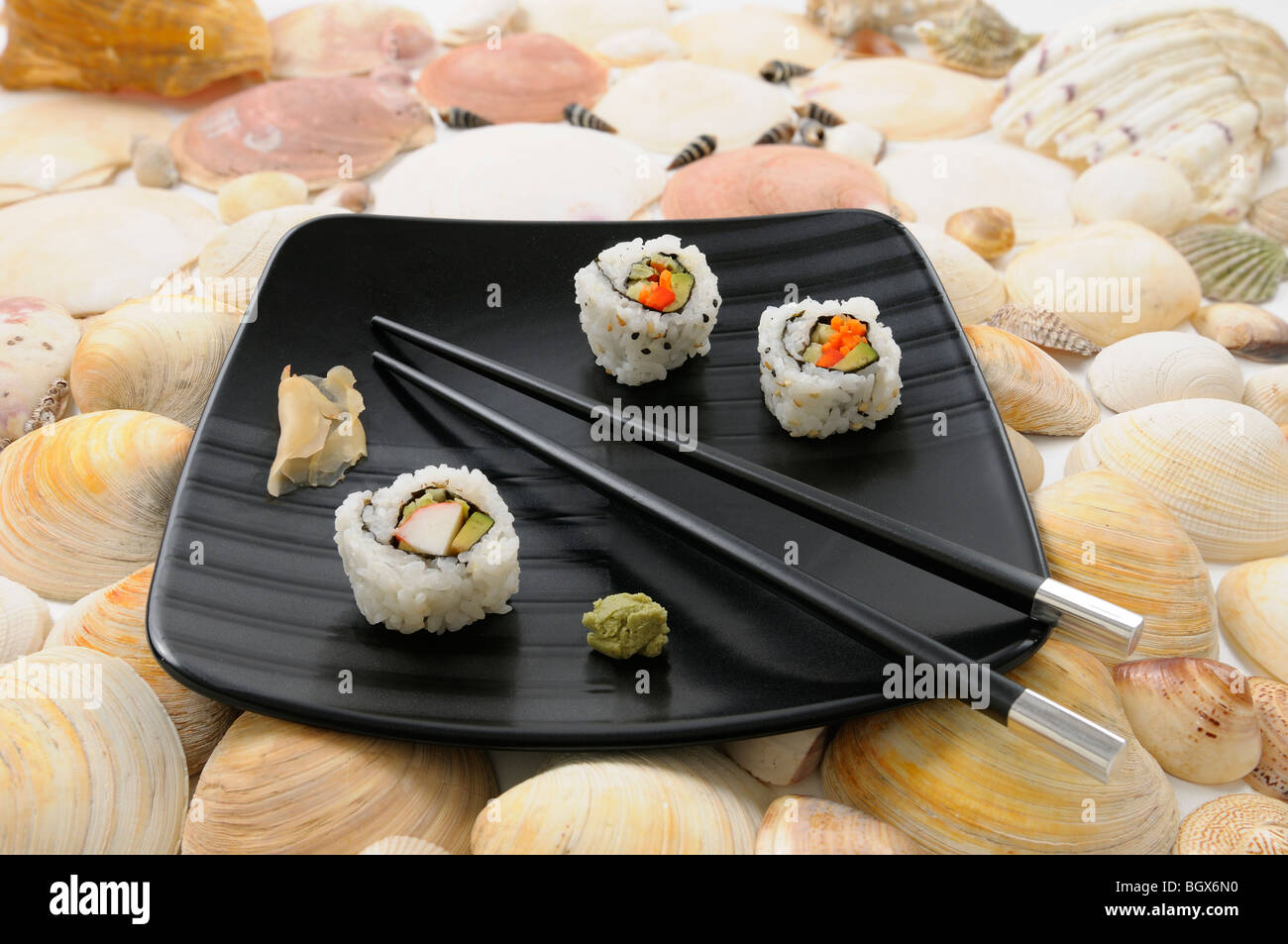 Japanische Krabben Uramaki Sushi und Stäbchen mit Wasabi und Ingwer auf einem schwarzen Teller umgeben von Muscheln Stockfoto